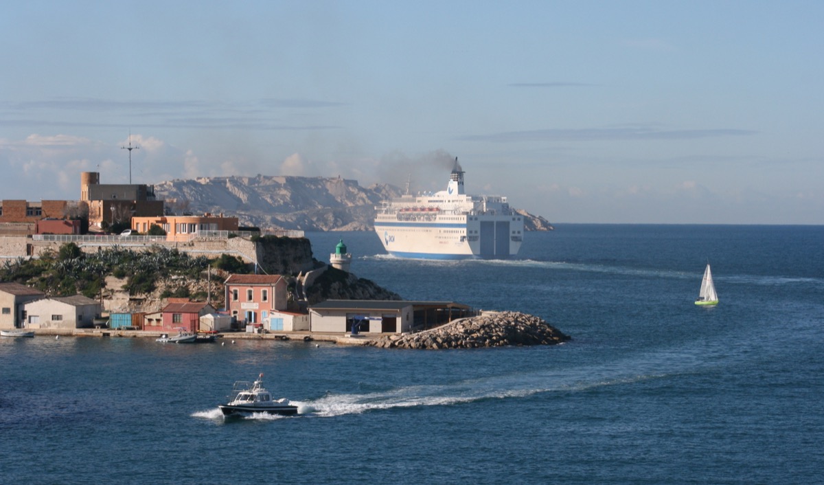 Eine große weiße Fähre verlässt den Hafen von Marseille und passiert eine felsige Halbinsel mit kleinem Leuchtturm